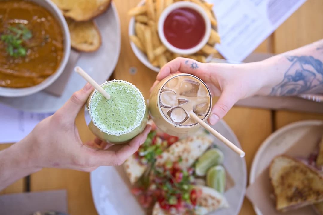 Two people raising iced drinks over a café table of food